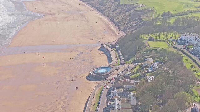 Filey Promenade tidal pool on Ravine Road Filey North Yorkshire England with circular seawater pool, curved sea wall and wide sandy beach along North Sea coast, drone orbit shot
