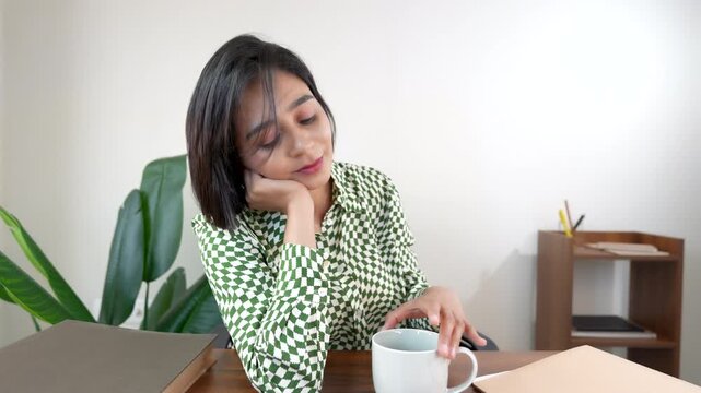 Close up of happy thoughtful beautiful young Asian business woman sits on chair at workplace desk hold coffee cup thinking pondering creative plan.