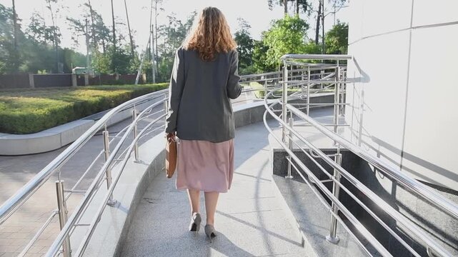Slow motion back view of a young blonde business woman walking up a ramp toward an office building entrance, carrying a coffee and a bag with documents. 