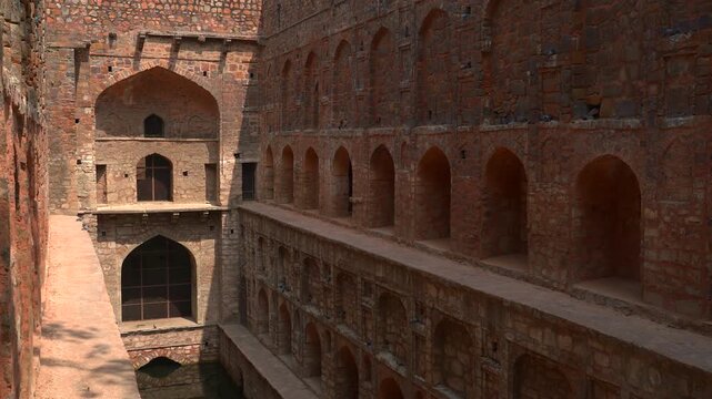 A wide static shot of the Agrasen ki Baoli, a 14th-century historical stepwell in New Delhi, featuring its unique red stone walls with multiple levels of arched niches and a central well.