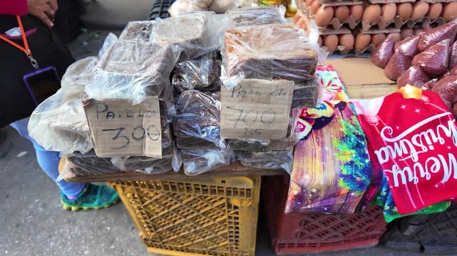 Close-up slow motion street vendor selling panela, traditional unrefined sugar cane blocks, packaged in plastic bags at local market. Handwritten price signs are visible as person arranges products