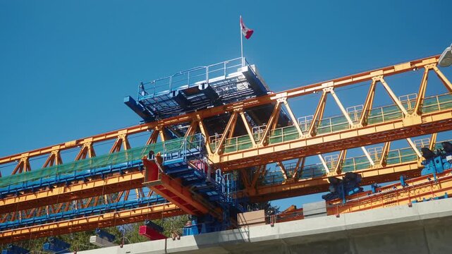 static Handheld perspective showing the detailed steel truss structure of a yellow and blue segment launcher with a Canadian flag waving overhead during the Surrey Langley SkyTrain construction