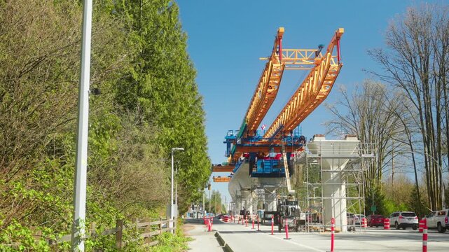 Stationary wide angle perspective from street level shows the massive yellow segment launcher extending over the road to construct the elevated concrete transit guideway for Surrey Langley SkyTrain