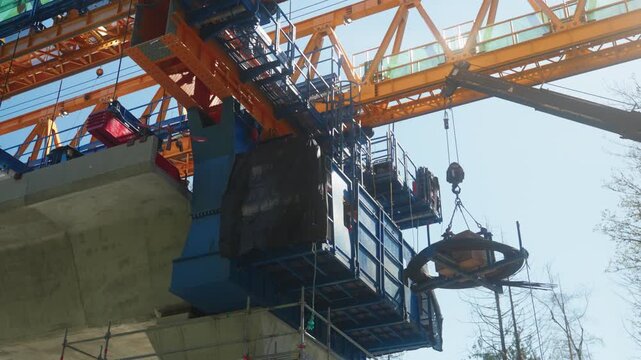 Handheld perspective looks up as a crane hoists a heavy coil of steel materials next to the blue support structures of a massive yellow segment launcher while the sun flares through the machinery