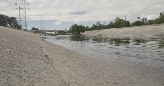 Cinematic daytime 4K 10-bit footage captured from the riverbank of the LA River looking downstream toward a bridge structure as the water flows steadily through the channel.