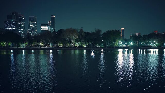 Lake with skyline reflections and illuminated skyscrapers in Benchakitti park at night, Urban waterfront with city lights and modern highrise buildings, Bangkok Thailand 4k