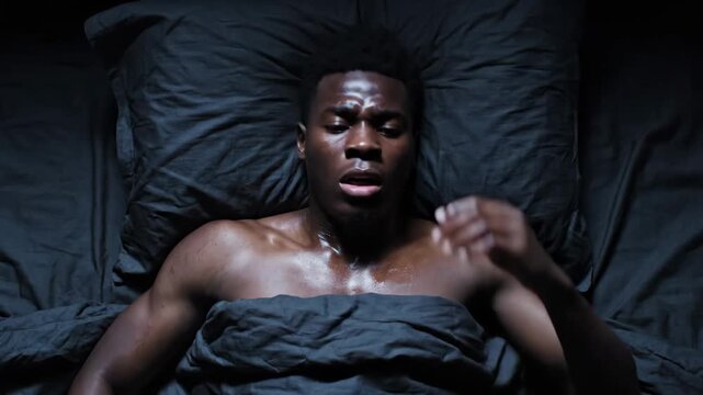 Overhead shot of a young man waking up in a dark bedroom, sweating and distressed from a nightmare. He looks around in fear, then slowly calms down, reflecting on the disturbing dream.