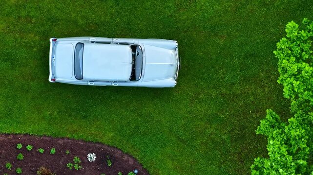 Cinematic aerial camera movement over a light blue oldtimer parked in a lush, green garden.