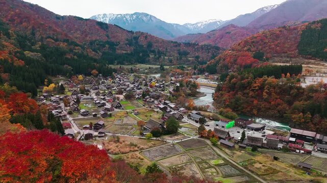 Panoramic drone shot of the historic Shirakawa-go village during autumn. Traditional thatched-roof farmhouses are surrounded by vibrant red and orange foliage with mountains in the background.