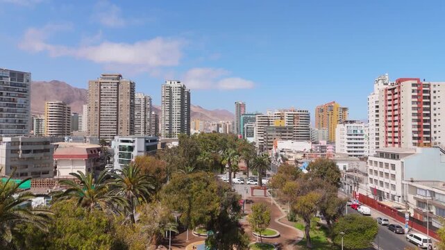 Second part of the aerial drone flyover of Parque Brasil in Antofagasta, Chile. Smooth forward motion over treetops revealing more of the coastal city skyline and surrounding desert mountains.
