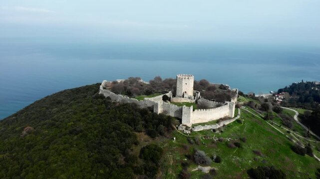 A hilltop castle with stone walls and a central tower, surrounded by lush greenery and overlooking the expansive sea. Suitable for historical documentaries or travel content.
