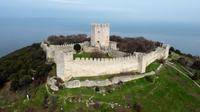 A stone fortress with a central tower on a green hilltop by the sea. Ideal for historical documentaries or travel content.