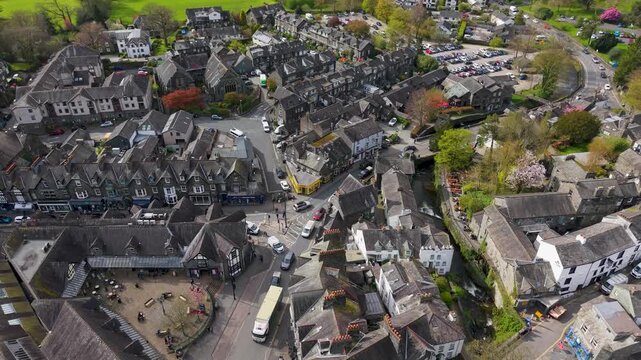 A591 Ambleside reverse rising drone shot pulls back and climbs above slate-roof Lakeland buildings, busy junction and cars, revealing the town layout in Cumbria, England.