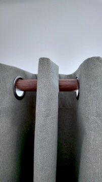 Grey fabric curtains with eyelets hanging from a dark wood curtain rod. vertical video