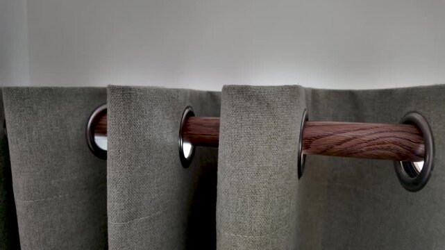 Grey fabric curtains with eyelets hanging from a dark wood curtain rod. dolly left