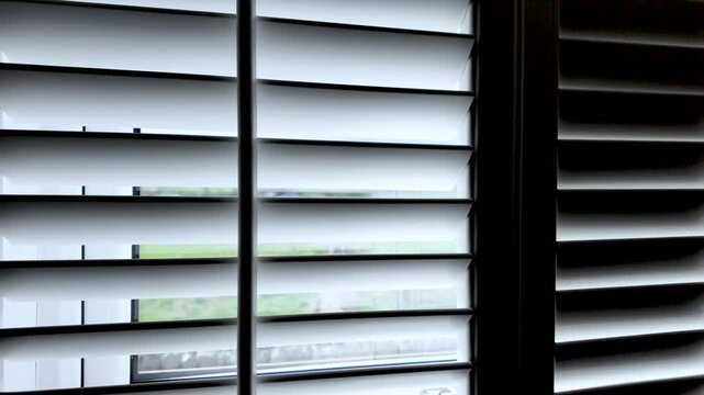 White louvered shutters partially closed, filtering light from a window. close up, dolly right