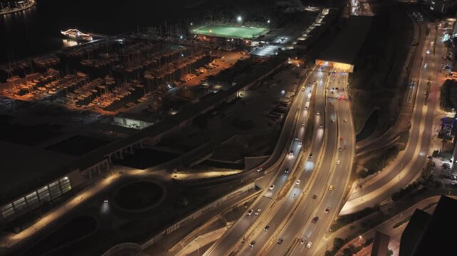 Beautiful aerial perspective of a bustling coastal city highway at night, with car headlights illuminating the road next to a tranquil marina filled with docked yachts and a lit sports field