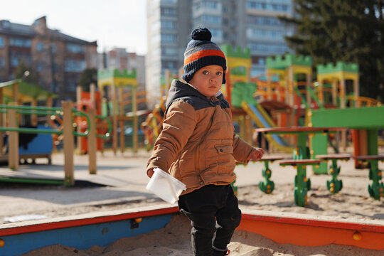 A young child stands in a sandbox at a park during daytime. The child holds paper and looks around while wearing a winter coat and hat