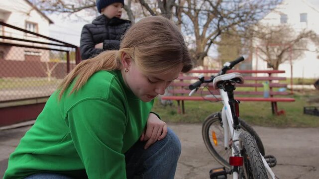 Focused teenage girl in casual clothes inflating a bicycle tire with a manual pump in a park, preparing her bike for a ride while a boy watches in the background on a spring day