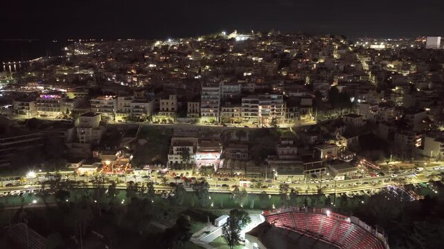 Aerial perspective showing the illuminated city of piraeus at night, with bustling traffic along the coast, the veakeio theatre, and the surrounding residential buildings of kastella hill