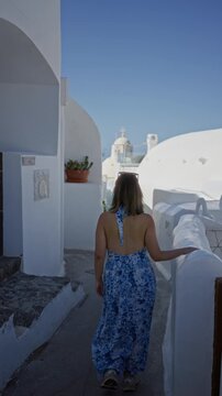 Woman walking with hand on railing, bare back visible, through a narrow white santorini street with domed buildings; serenity travel.