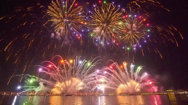 Dramatic grand finale fireworks display exploding in vibrant colors over a calm river with urban city lights reflecting on the water at night.
