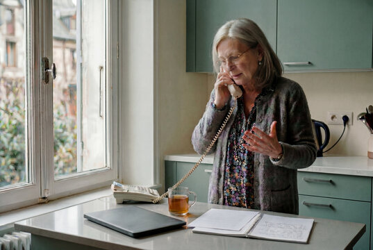 Seniorin im Gespr&auml;ch am Schnurtelefon in heller K&uuml;che mit Notizbuch und Teetasse am Fensterplatz