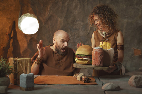 Happy prehistoric couple having lunch together in their cave: they are eating fast food