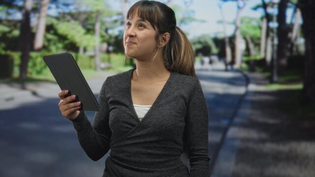 Woman holding a tablet and reading on a tree lined park path, smiling and looking at screen with casual posture; contentment.