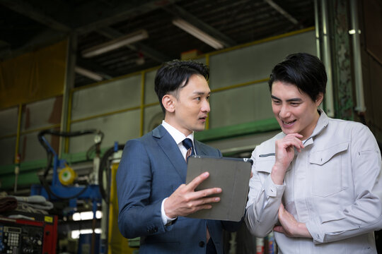 製造業の工場でタブレットを持つ作業員と信金などの打ち合わせをする営業ビジネスマンの2人　設備投資で業務効率などの借り入れのイメージ　左にコピースペースあり