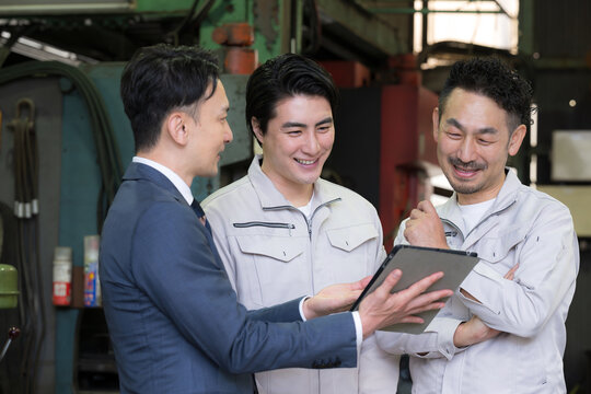 工場でタブレットを使い３人で打ち合わせする作業員とビジネスマンの現場、dxやIT、AI化の機械の導入や信金ビジネスマンが借り入れ提案のイメージ