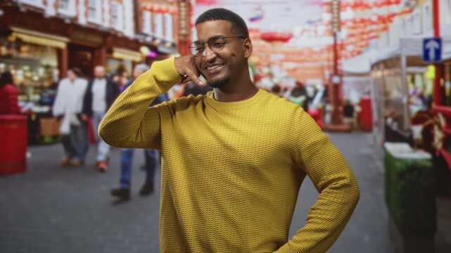 Young african man with hand to ear making a phone call gesture on a crowded street wearing yellow sweater and glasses; connection joy.