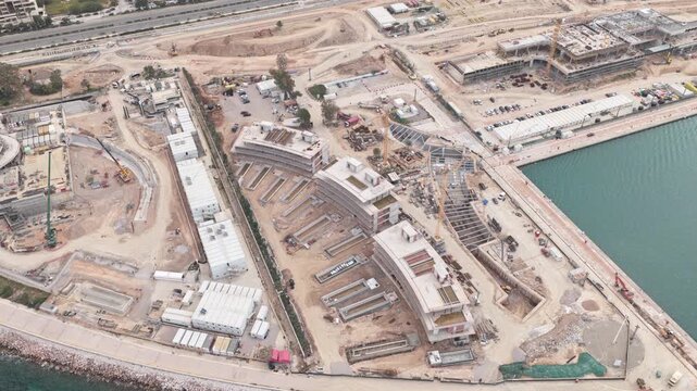 High-angle drone footage showing the development of a large waterfront construction project with several buildings under construction, heavy machinery, and foundational work near the sea