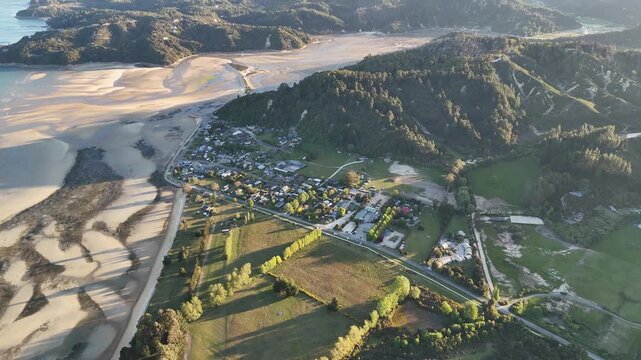 Aerial sunset view of Marahau in Abel Tasman region, South Island New Zealand, showing coastal settlement, rural farmland and tidal flats surrounded by forested hills in warm golden light