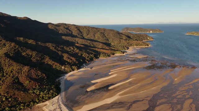 Aerial sunset view of Abel Tasman coastline near Marahau, New Zealand. Sweeping tidal sandbanks, rugged forested hills and calm blue ocean in warm golden hour light. Wide shot.