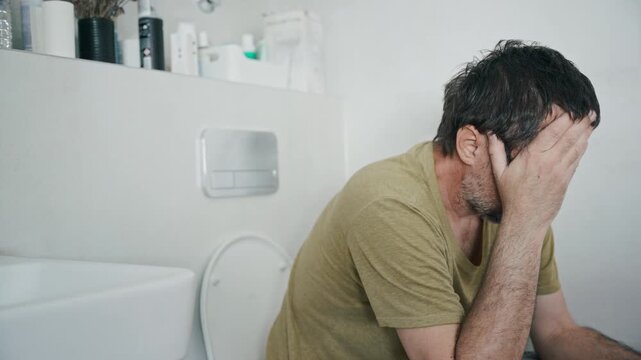 A distressed man sits on a toilet in a bathroom, covering his face with his hand while experiencing pain or nausea. The image conveys illness, hangover, stress or constipation. 4K.