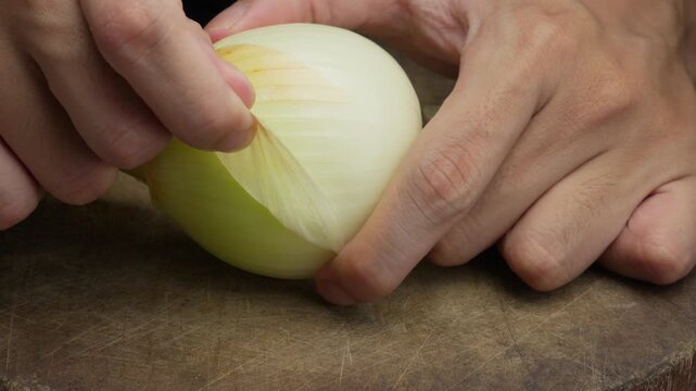 A close up shot of a person's hands carefully peeling the dry outer skin off a fresh white onion on a wooden cutting board in a kitchen