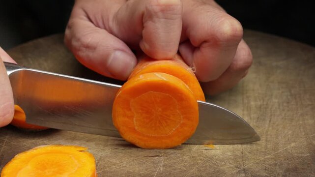 A close up shot of a chef's hands using a sharp knife to slice a fresh carrot into rounds on a wooden cutting board. Ideal for cooking and nutrition content.