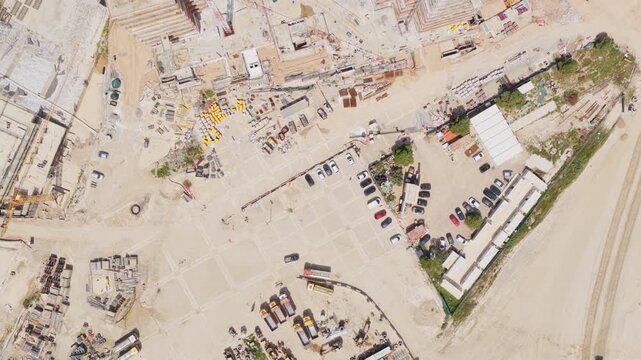 High aerial top-down perspective showing the development of a large building foundation, with heavy machinery, materials, and a tower crane shadow moving as the camera slowly pulls away