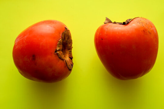 Two juicy orange persimmons lie on a bright lemon-yellow background, forming an energetic and warm color composition. The smooth skin of the fruits softly reflects the light, highlighting their natura