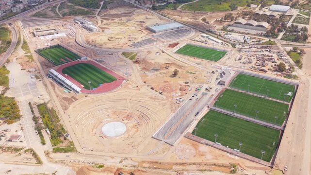 Expansive aerial perspective showing the ongoing construction of a modern sports complex, featuring multiple football fields, an athletics track, and an amphitheater surrounded by earthworks