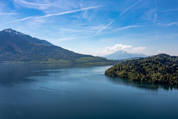 Fototapeta premium Tranquil aerial view of Lake Zug shoreline - forested peninsula and distant Alps under blue sky