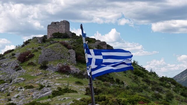 Aerial shot of a Greek flag and ancient hilltop stone ruins on a rocky slope.