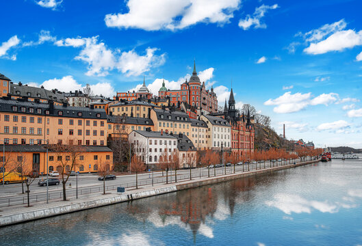 A scenic view of the historic colorful buildings along the Sodermalm waterfront in Stockholm, Sweden, reflected in the calm waters of Lake Malaren under a blue sky.