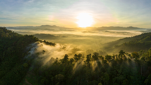 Breathtaking aerial view of the sun rising over a mountain valley covered in a dense sea of rolling clouds. The golden light illuminates the mist, creating a dramatic and ethereal atmosphere at dawn