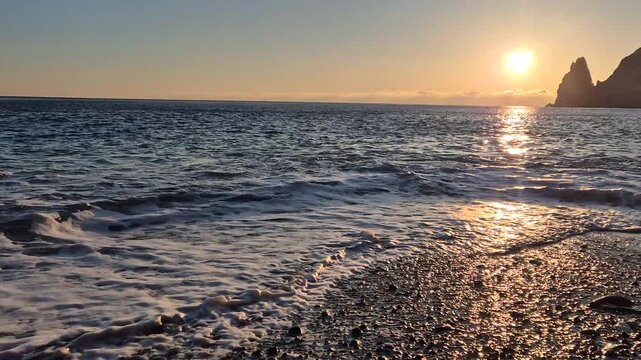 Sunset beach waves rolling across a pebbly shore during a golden sunset with dramatic cliffs and a serene ocean horizon
