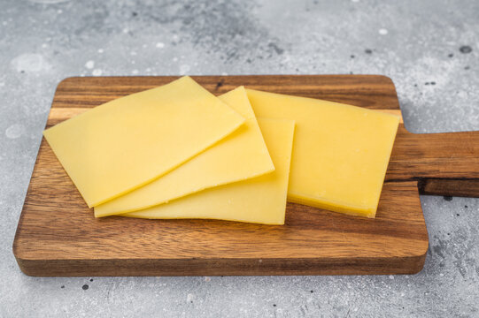 Stack of semi hard yellow cheese slices on rustic wooden cutting board, copy space on gray stone background, dairy ingredient