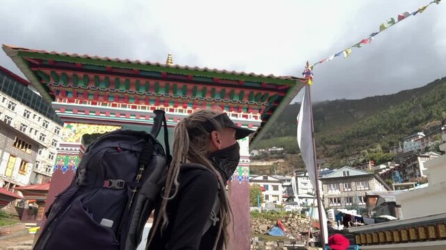 Female traveler with dreadlocks and backpack filming vlog while walking to Namche Bazaar, Everest Base Camp trek, Nepal. Himalayan village, prayer flags, travel adventure journey