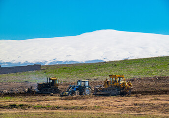 Fototapeta premium Tractors working in field with snowy mountain backdrop