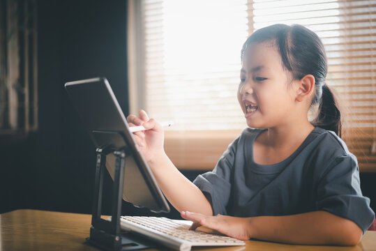 Happy young Asian girl using tablet with keyboard and stylus for online learning, little student smiling during digital classroom session at home office workspace.
